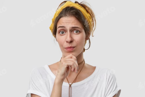 Preview: Indecisive pretty woman holds chin, looks with clueless expression at camera, wears yellow headband,