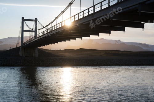 Preview: Suspension bridge over river at sunset with mountain backdrop.