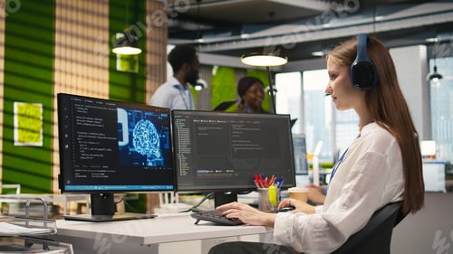 Preview: Young female programmer in office reviewing AI system metrics, listening music