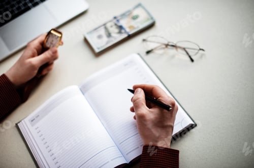 Preview: Candid authentic man writes a business plan in notebook holding a watch in the other hand at home