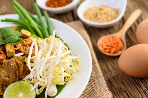 Preview: Pad Thai in a white plate with lemon, eggs and seasoning on a wooden table.