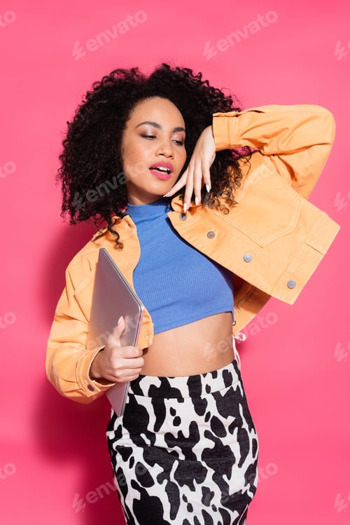 Preview: stylish african american woman posing with laptop on pink
