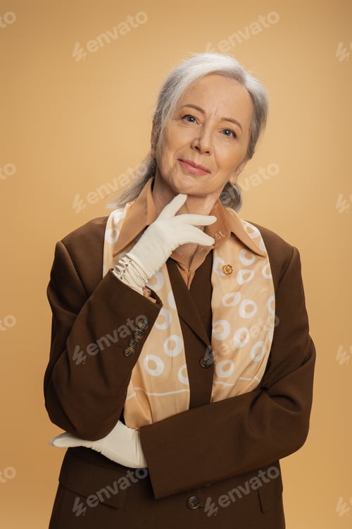 Preview: portrait of pleased senior woman in white gloves and brown suit looking at camera isolated on beige