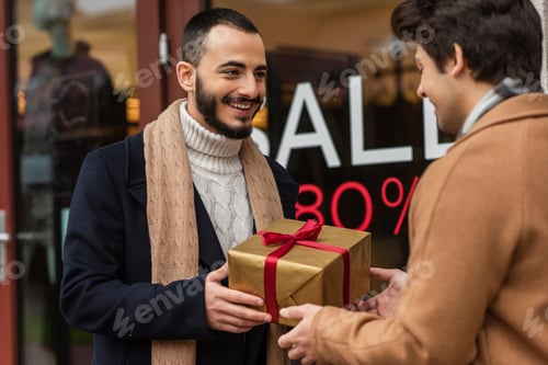 Preview: happy bearded man holding Christmas present near blurred boyfriend and showcase with sale lettering