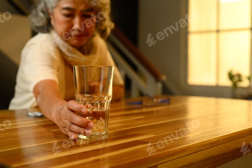 Preview: Senior woman reaching for a glass of water to take a pill at kitchen counter