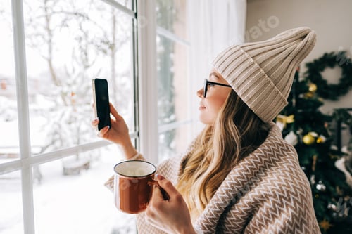 Preview: Young caucasian woman standing near window at home and taking photo after snowfall in the morning.