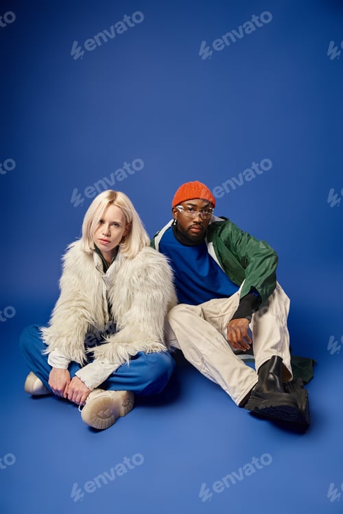 Preview: stylish african american man and blonde woman in winter attire sitting together on blue backdrop