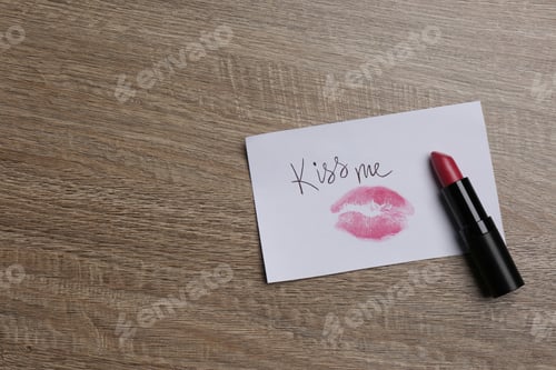 Preview: Paper card with lip print, phrase Kiss Me and lipstick on wooden table, top view. Space for text
