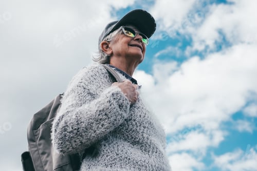 Preview: Smiling active senior woman with cap walking outdoors holding backpack looking at blue sky