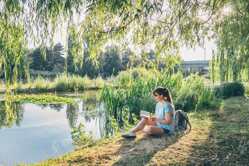 Preview: Young woman reading book by lake in peaceful natural setting