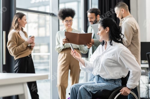 Preview: happy disabled businesswoman in wheelchair holding smartphone near blurred multiethnic colleagues
