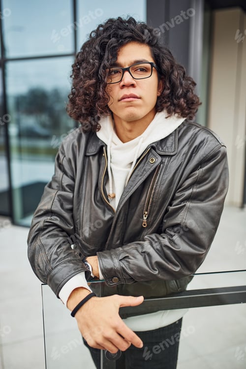 Preview: Handsome young man with curly black hair and in glasses is on the street against building