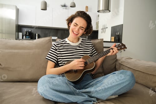 Preview: Portrait of cheerful young woman playing her ukulele, singing and laughing, sitting in living room