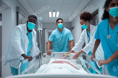Preview: Mixed race doctors wearing face masks transporting patient in hospital bed
