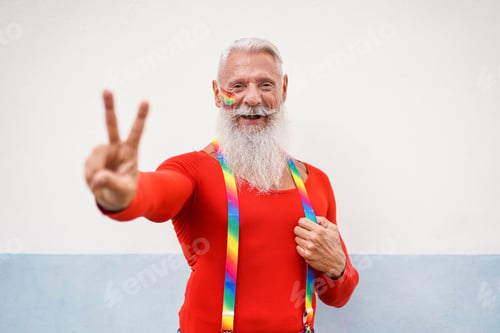 Preview: Cheerful hipster senior man at gay pride event doing peace gesture with hand