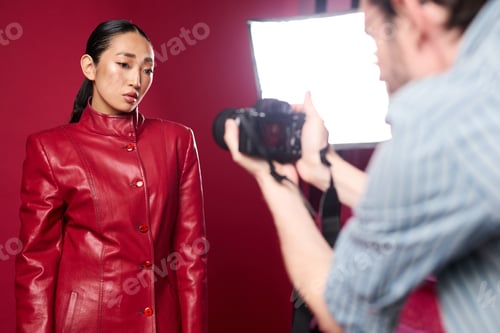 Preview: Fashionable asian woman in red leather jacket posing in front of a red wall with a man taking her