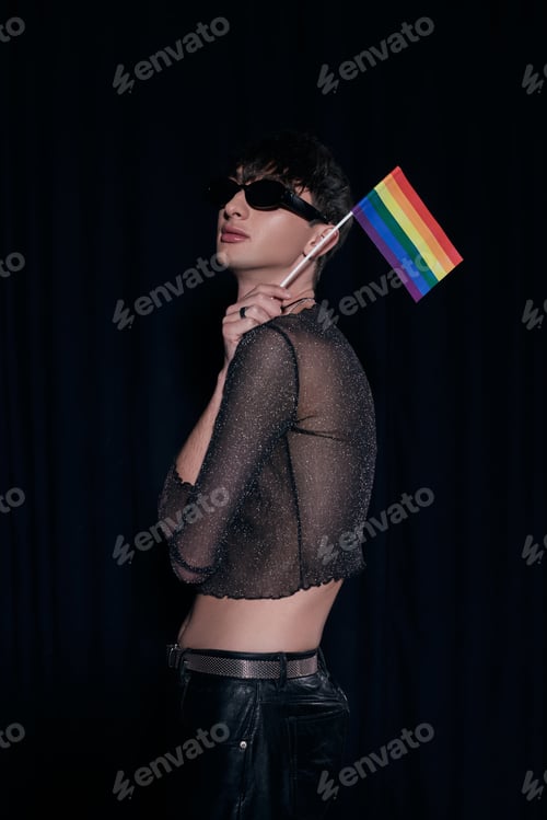 Preview: Fashionable and young homosexual man in sunglasses and shiny top holding rainbow flag during lgbt