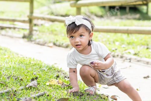 Preview: little brunette girl, on the side of the road leaning over, touching the grass while looking up.