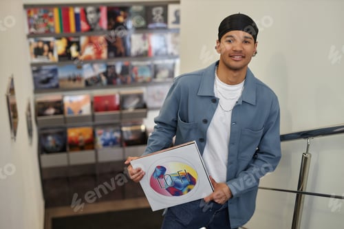 Preview: Smiling black man holding vinyl record standing on stairs in music store