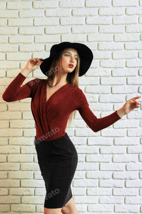 Preview: Model poses in a studio wearing a red top and black skirt with a black hat against a white wall