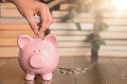Preview: Woman’s hand dropping a coin into piggy bank with stack of books in the background