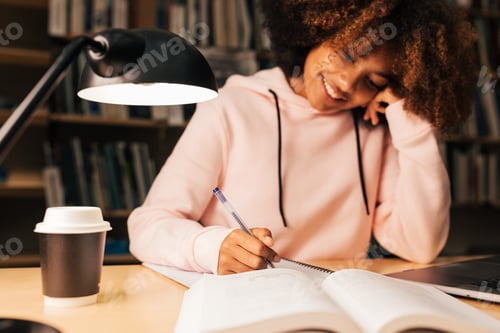 Preview: Cheerful student writing on notebook while sitting at desk in library