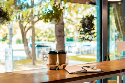 Preview: two coffee in paper cups, open notebook with pen on cafe counter