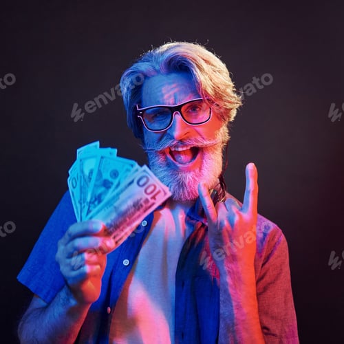 Preview: Holding cash. Neon lighting. Stylish modern senior man with gray hair and beard is indoors
