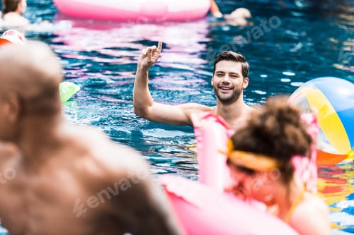 Preview: smiling man with raised finger in swimming pool talking to friends