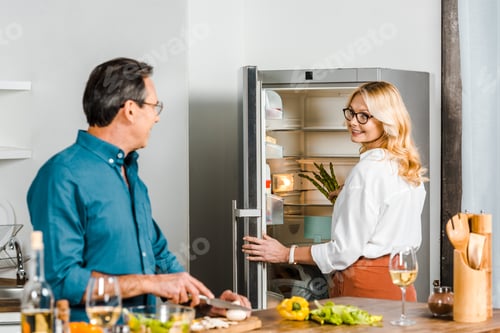 Preview: mature wife taking asparagus from fridge and husband cutting vegetables in kitchen