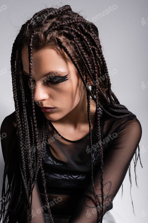 Preview: brunette woman with braids posing in futuristic makeup and black clothes isolated on grey