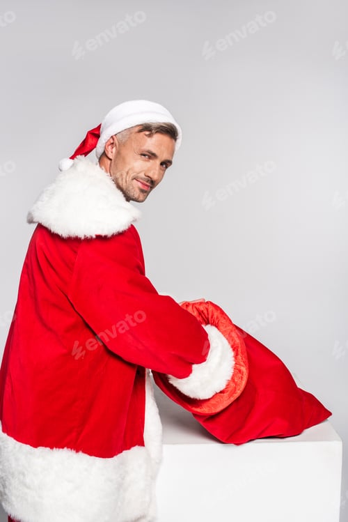 Preview: handsome man in santa costume holding red bag and smiling at camera isolated on grey