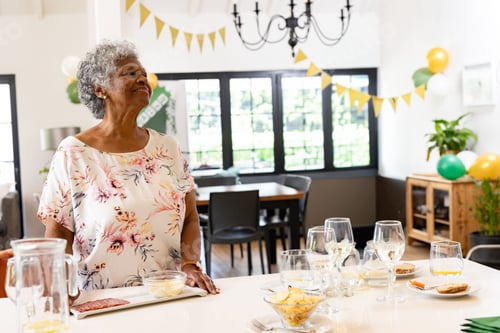 Preview: Senior African American woman standing at island in dining area with wine glasses banners balloons