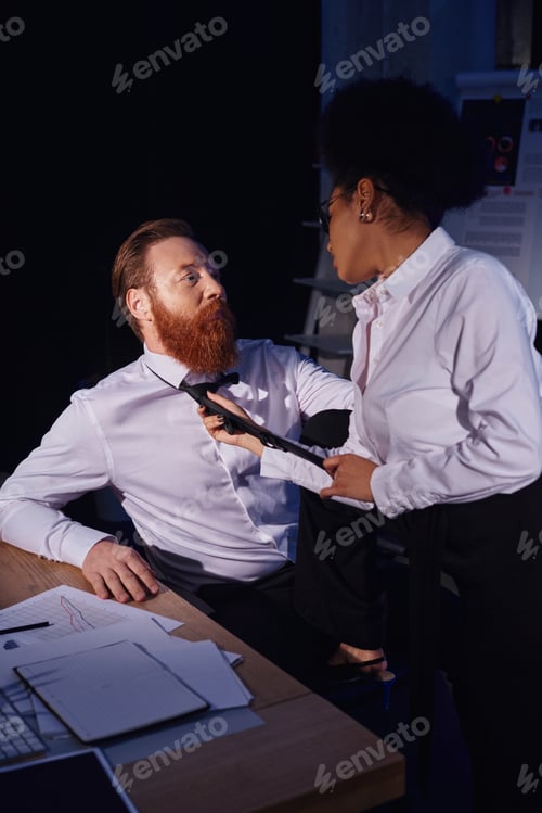 Preview: african american woman pulling tie of bearded businessman in night office, love affair at work
