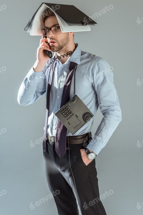 Preview: Confused businessman holding notebook on his head while talking on phone isolated on grey