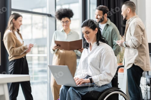 Preview: disabled businesswoman in wheelchair using laptop near blurred multiethnic colleagues, inclusion