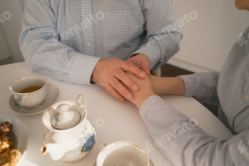 Preview: Elderly couple holding hands together while enjoying tea and cookies at home