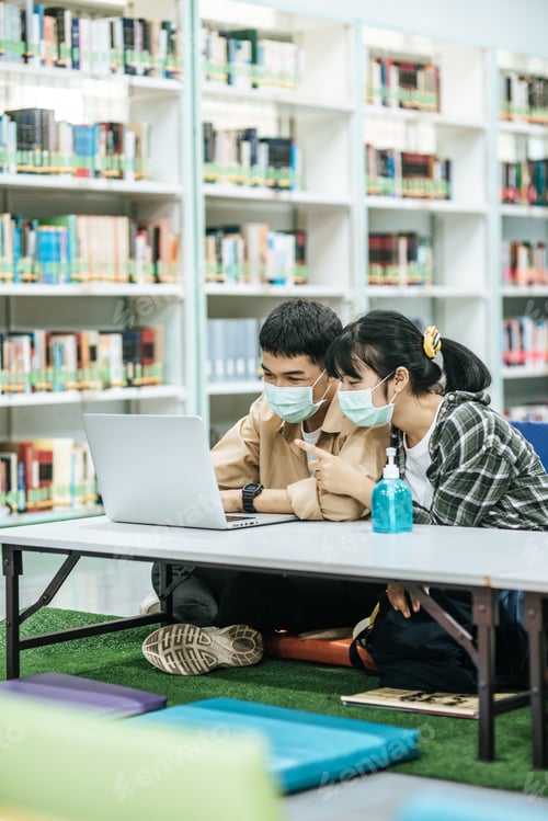 Preview: Men and women wear masks and use a laptop to search for books in the library.