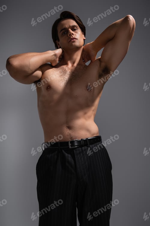 Preview: good looking man with muscular body posing in black trousers and looking at camera on grey