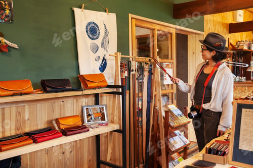 Vista previa: Japanese woman wearing hat and glasses browsing merchandise in a leather shop.