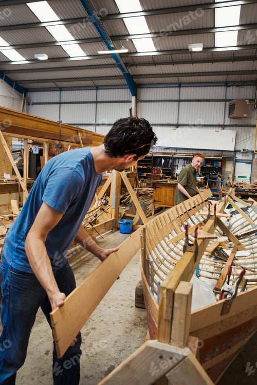 Preview: Two men in a boat-builder's workshop, working together on a wooden boat hull.