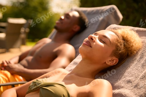 Preview: Couple On Holiday Wearing Swimming Costumes Sunbathing On Loungers By Hotel Swimming Pool
