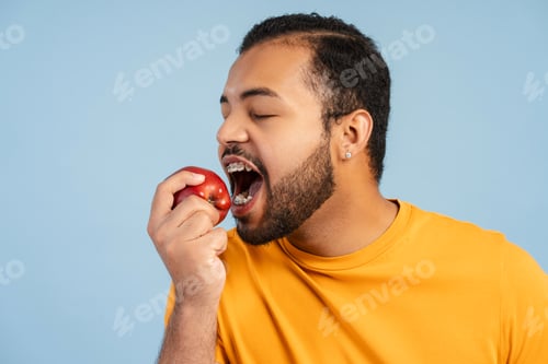 Preview: Happy african american man with braces in orange shirt holding and eating red apple isolated