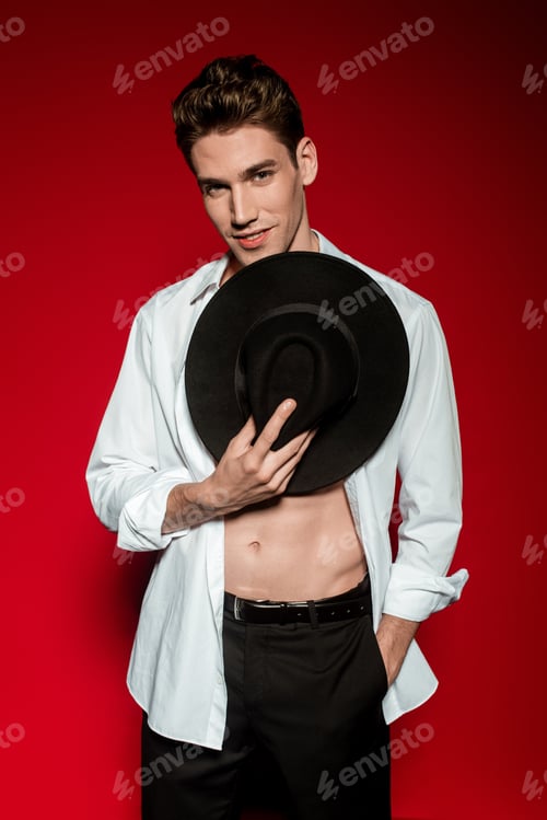Preview: sexy young elegant man in unbuttoned shirt with muscular bare torso and hand in pocket holding hat