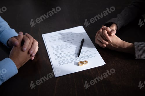 Preview: partial view of couple sitting at table with clenched hands near divorce documents and rings
