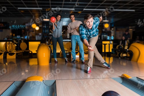 Preview: smiling young man throwing bowling bowl on skittle alley near multicultural friends