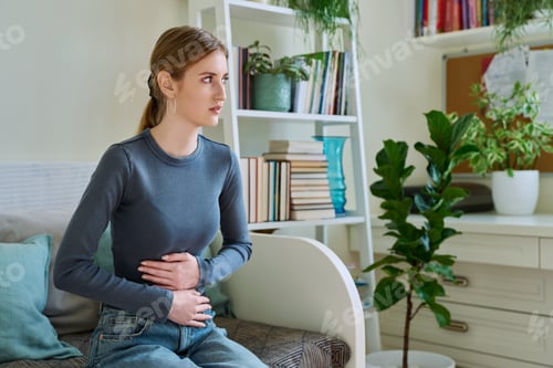 Preview: Young teenage female experiencing abdominal pain, sitting on couch at home