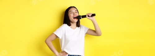 Preview: Happy asian woman singing song in karaoke, holding microphone, standing over yellow background