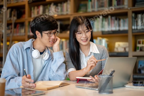 Preview: Glasses asian man student looking at laptop as woman friend pointing while sits at table in library.