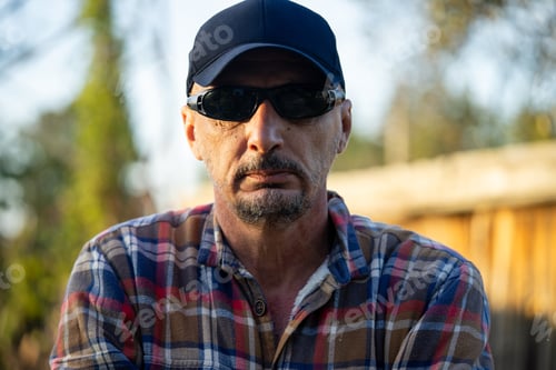 Preview: Self assured mature man in checkered shirt and straw hat crossing arms in countryside